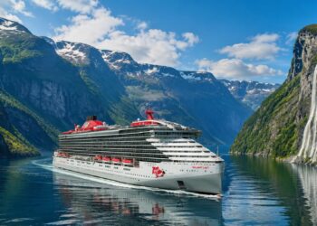 Virgin Voyages' Valiant Lady sailing through the Norwegian Fjords surrounded by mountains and waterfalls.