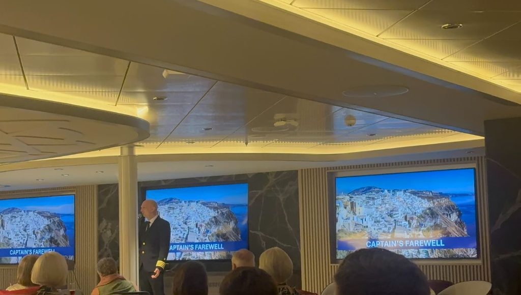 The Captain addressing guests during the Captain's Farewell on Douglas Mawson, with Santorini on the screens behind him