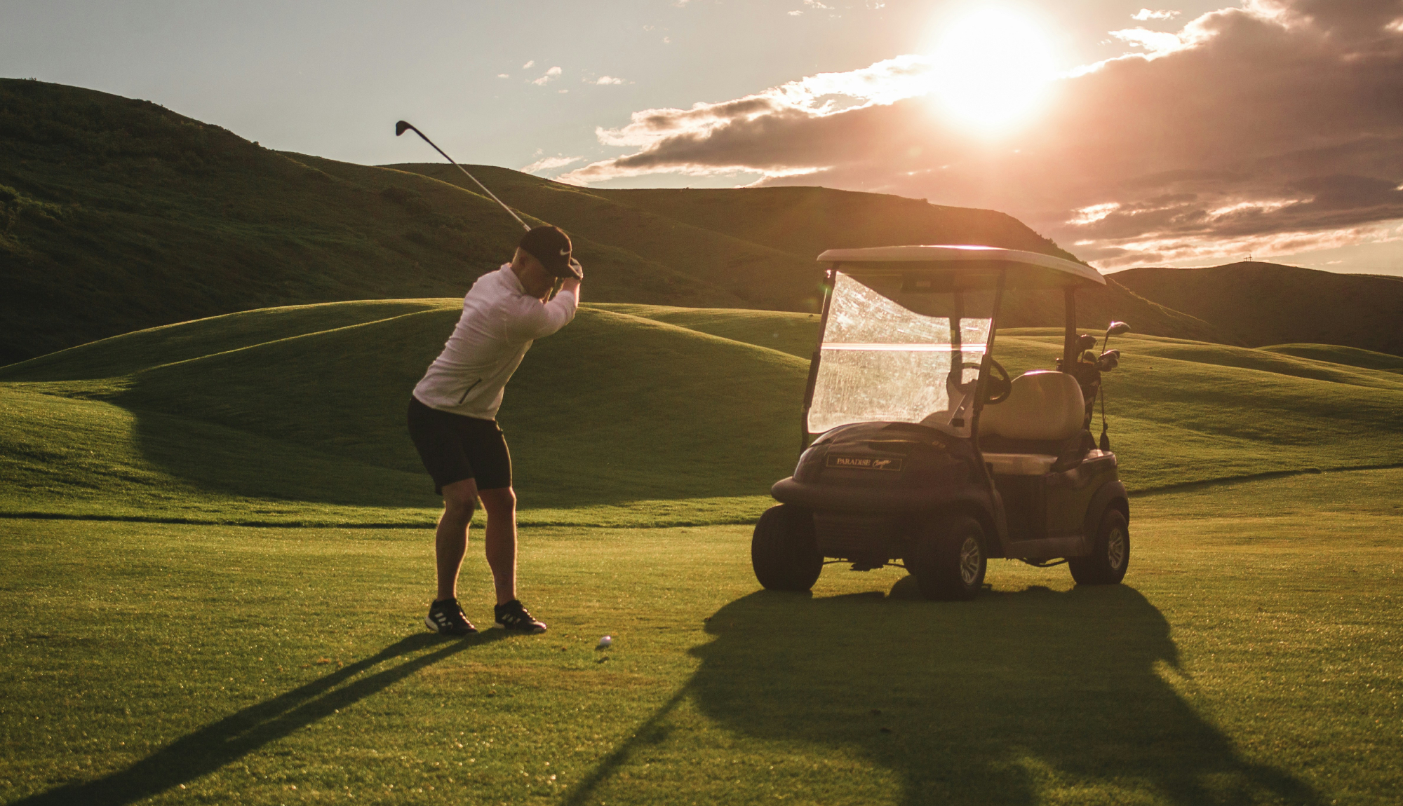 Man taking a swing on a golf course with a buggy behind him as part of an Azamara Golf Cruise booked via Paramount Cruises.