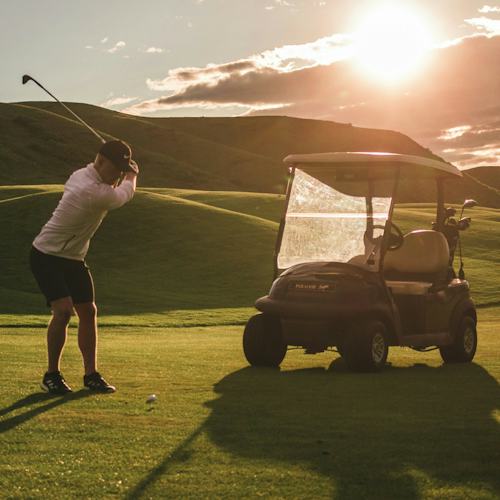 Man taking a swing on a golf course with a buggy behind him as part of an Azamara Golf Cruise booked via Paramount Cruises.